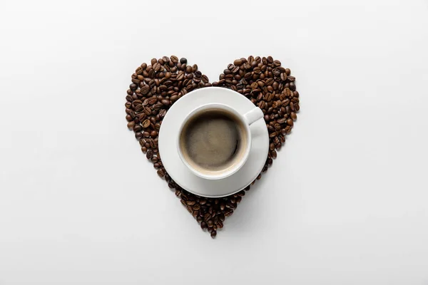 Vue de dessus de café savoureux dans une tasse sur soucoupe avec coeur en grains de café sur fond blanc — Photo de stock