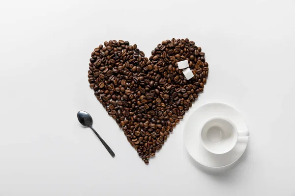 Vista dall'alto della tazza vuota vicino al cuore fatta di chicchi di caffè con zucchero su sfondo bianco — Foto stock