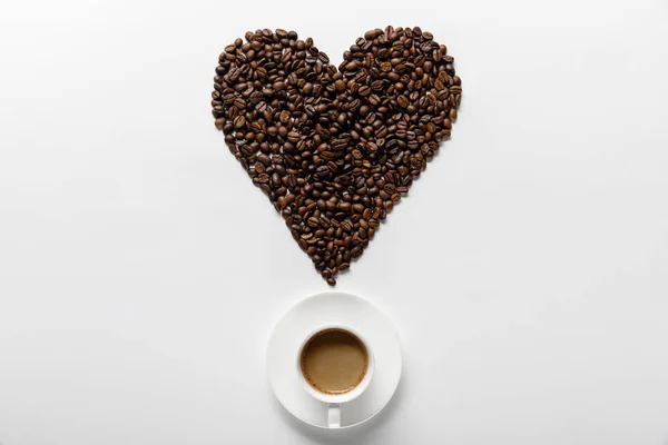 Vista dall'alto di delizioso caffè in tazza sul piattino con cuore fatto di grani di caffè su sfondo bianco — Foto stock