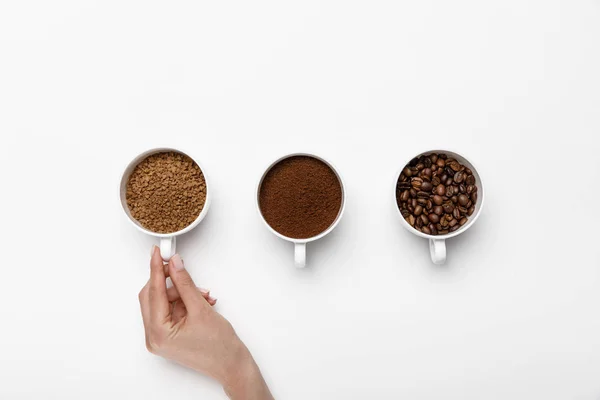 Vista recortada de la mano femenina cerca de tres tipos de molienda de café en tazas sobre fondo blanco - foto de stock