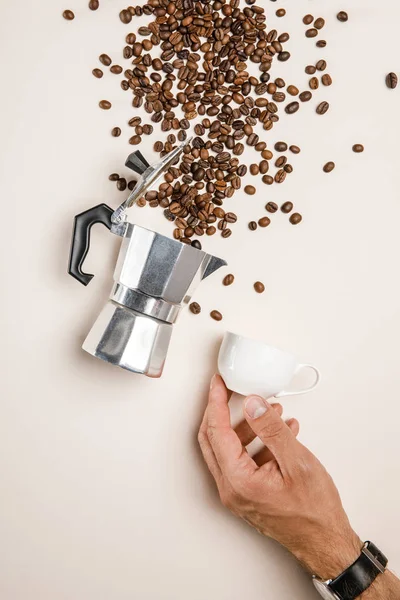 Vista parziale dell'uomo che tiene la tazza vicino alla caffettiera in alluminio e ai chicchi di caffè freschi sparsi su sfondo beige — Foto stock