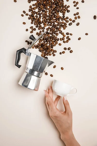 Partial view of woman holding cup near aluminium coffee pot and scattered fresh coffee beans on beige background — Stock Photo