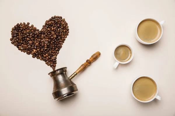 Top view of cups with coffee near heart made of coffee grains and cezve on beige background — Stock Photo