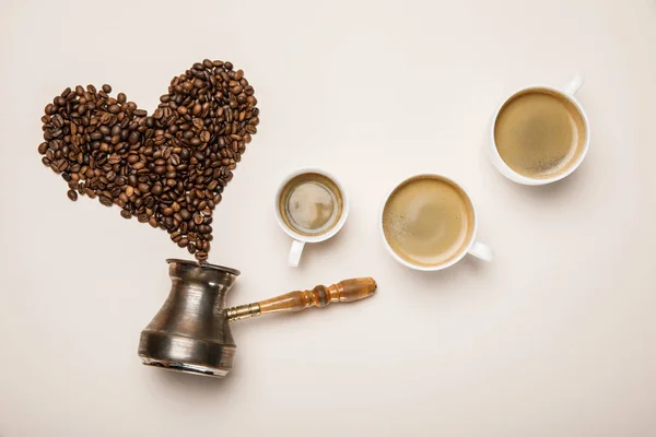 Draufsicht auf Tassen mit frischem Kaffee in Herznähe aus Kaffeekörnern und Cezve auf beigem Hintergrund — Stockfoto