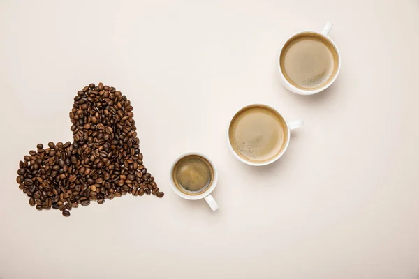 Draufsicht auf Tassen mit Kaffee in Herznähe aus Kaffeekörnern auf beigem Hintergrund — Stockfoto