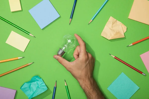 Cropped view of man holding light bulb near scattered pencils and crumpled paper on green — Stock Photo