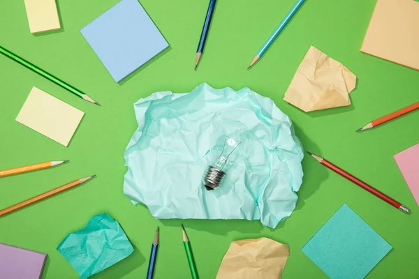 Верхний вид лампочки на большом куске скомканной бумаги рядом с разбросанной бумагой и карандашами на зеленом — стоковое фото
