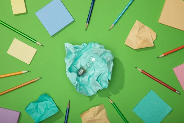 Vista superior da lâmpada no pedaço de papel amassado perto de papel espalhado e lápis em verde — Fotografia de Stock