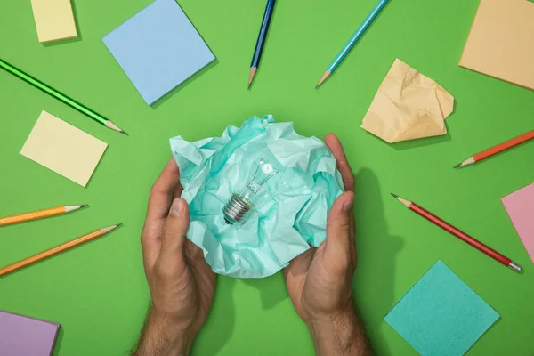 Abgeschnittene Ansicht eines Mannes, der zerknülltes Papier mit Glühbirne in der Nähe von Bleistiften und Papier auf Grün hält — Stockfoto