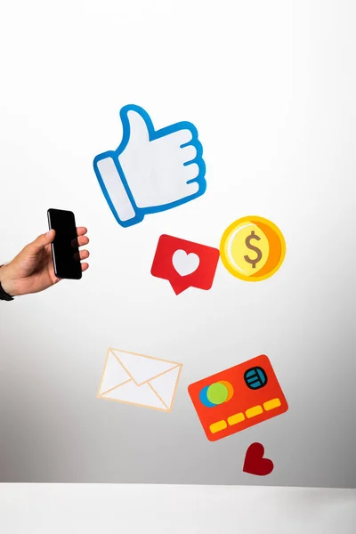 Cropped view of man holding smartphone near colorful paper icons on white and grey background — Stock Photo