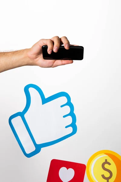 Cropped view of man holding smartphone above colorful paper icons with coin, heart and thumb up on white background — Stock Photo