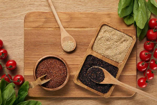 Top view of white, black and red quinoa in wooden bowl on chopping board near spinach leaves and tomatoes — Stock Photo