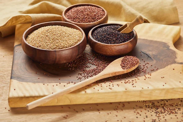White, black and red quinoa in wooden bowls and spoon on wooden chopping board near napkin — Stock Photo