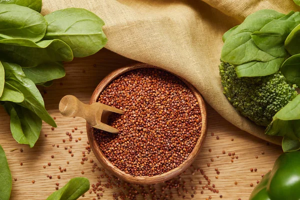 Top view of red quinoa in wooden bowl with spatula near beige napkin and green vegetables — Stock Photo