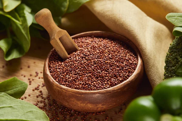 Vue rapprochée des graines de quinoa rouge dans un bol en bois avec spatule près de serviette beige et légumes verts — Photo de stock