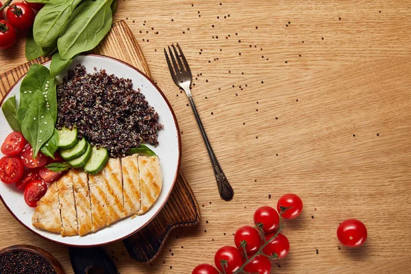 Vista superior de la quinua cocida con pechuga de pollo a la parrilla y verduras en plato blanco sobre mesa de madera con tenedor - foto de stock