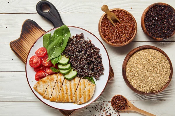 Blick von oben auf gekochte Quinoa mit gegrillter Hühnerbrust und Gemüse auf weißem Holztisch mit rohen, trockenen Samen in Schalen — Stockfoto