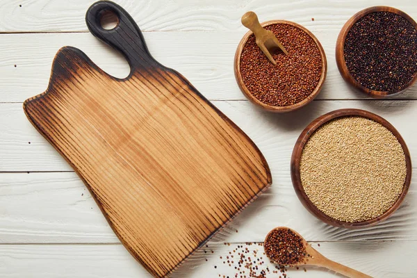Vue du dessus du quinoa blanc, noir et rouge dans des bols en bois près de la planche à découper sur table blanche — Photo de stock