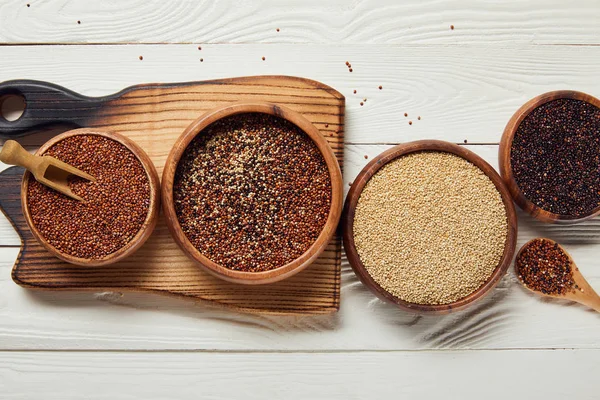 Top view of white, black and red quinoa seeds in wooden bowls on white table with chopping board — Stock Photo