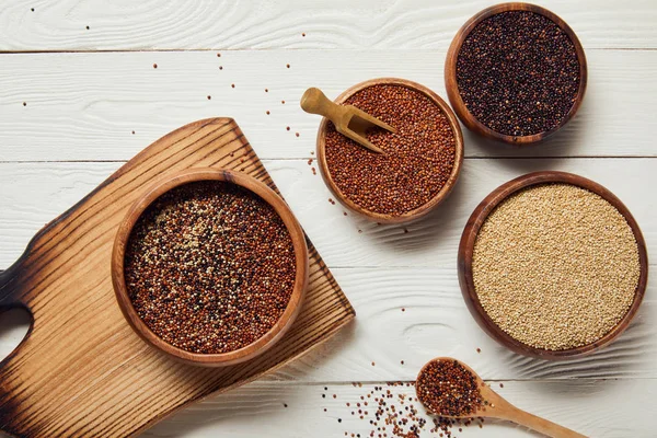 Vue du dessus des graines de quinoa crues blanches, noires et rouges dans des bols en bois sur table blanche avec planche à découper — Photo de stock