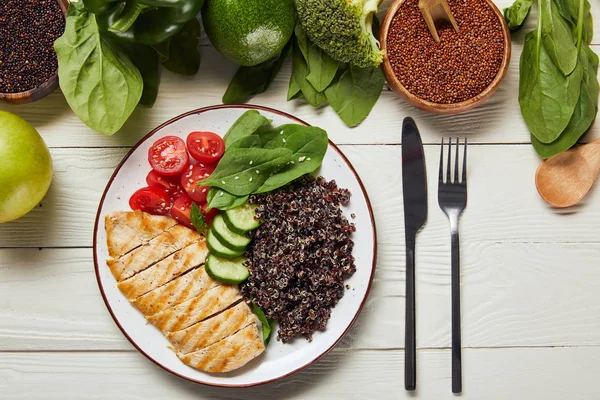 Vista superior de la deliciosa quinua servida con pechuga de pollo a la parrilla y verduras en plato blanco sobre mesa de madera - foto de stock