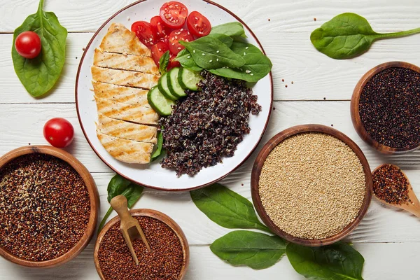 Top view of cooked quinoa with grilled chicken breast and vegetables on white plate on wooden table with spinach leaves and raw seeds — Stock Photo