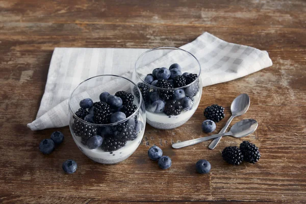 Gustosa colazione con yogurt e bacche in bicchieri vicino a cucchiaini e tovagliolo di stoffa sul tavolo di legno — Foto stock