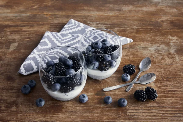 Gustosa colazione con yogurt e bacche in bicchieri vicino a cucchiaini e tovagliolo strutturato su tavolo di legno — Foto stock