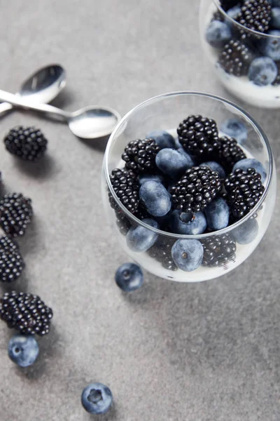 Messa a fuoco selettiva di gustosi yogurt con semi di chia, mirtilli e more in bicchieri vicino a cucchiaini e bacche sparse sulla superficie di marmo — Foto stock