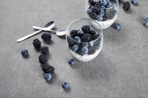 Вкусный йогурт с семенами чиа, черникой и ежевикой в стаканах возле чайных ложек и разбросанными ягодами на мраморной поверхности — стоковое фото