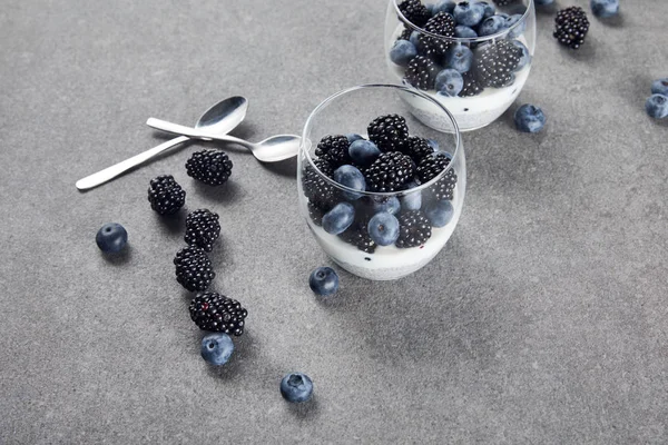 Iogurte saboroso com sementes de chia, mirtilos e amoras-pretas em copos perto de colheres de chá e bagas espalhadas na superfície de mármore — Fotografia de Stock