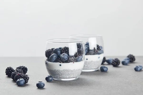 Selektiver Fokus von leckerem Joghurt mit Chiasamen, Blaubeeren und Brombeeren in Gläsern nahe verstreuter Beeren auf Marmoroberfläche isoliert auf grau — Stockfoto