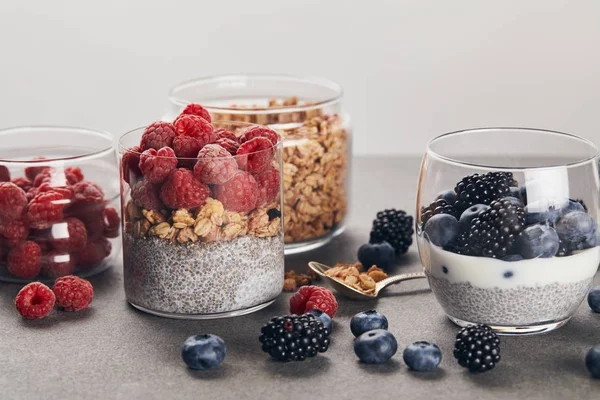 Vasos con yogur, bayas y copos de avena cerca de bayas dispersas en la superficie de mármol aislado en gris - foto de stock