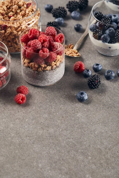 Yogurt con semi di chia, fiocchi d'avena e lamponi vicino a cucchiaino e vetro con mirtilli, more e yogurt sulla superficie di marmo — Foto stock