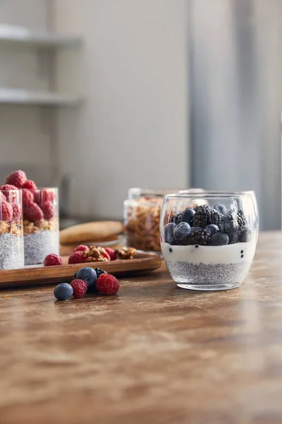 Enfoque selectivo de vasos con yogur, copos de avena y bayas en bandeja de madera en la mesa en la cocina - foto de stock