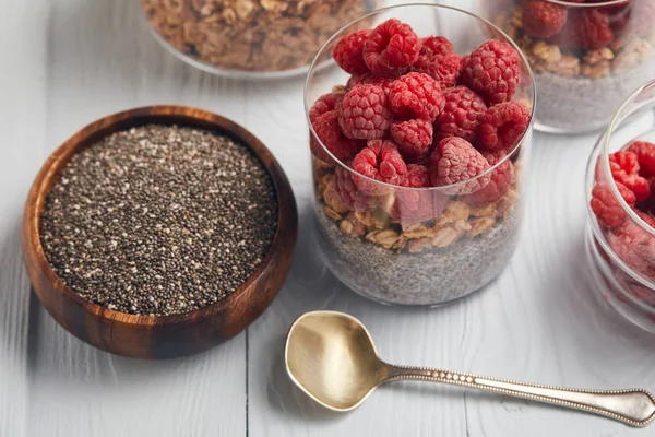 Glasses with yogurt, oat flakes and raspberries near wooden bowl with chia seeds on white table — Stock Photo