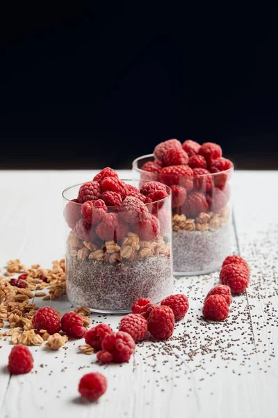 Glasses with yogurt, oat flakes and raspberries near scattered chia seeds, oat flakes and berries on white wooden table isolated on black — Stock Photo