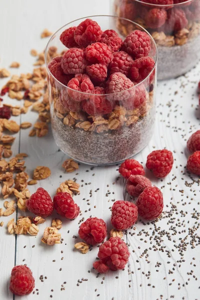 Selektiver Fokus von Gläsern mit Joghurt, Haferflocken und Himbeeren in der Nähe verstreuter Chiasamen, Haferflocken und Beeren auf weißem Holztisch — Stockfoto