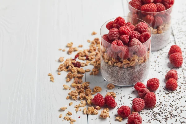 Glasses with tasty yogurt, oat flakes and raspberries near scattered chia seeds, oat flakes and berries on white wooden table — Stock Photo