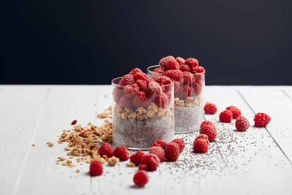 Iogurte saboroso com sementes de chia, flocos de aveia e framboesas em copos perto de sementes espalhadas, flocos de aveia e bagas na mesa de madeira branca isolada em preto — Fotografia de Stock