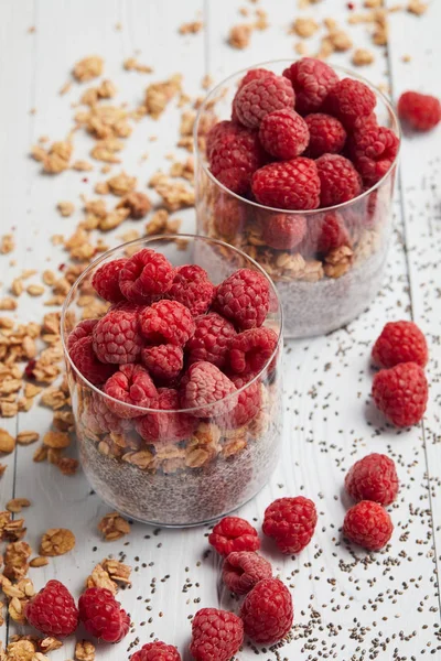 Foco seletivo de framboesas, flocos de aveia e iogurte com sementes de chia em copos perto de aveia espalhada, bagas e sementes em mesa de madeira branca — Fotografia de Stock