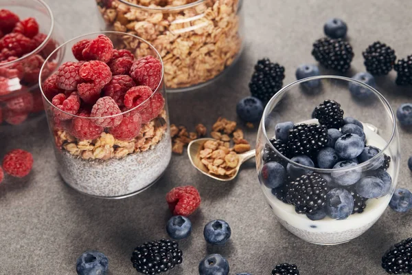 Selektiver Fokus von leckerem Joghurt mit Chiasamen, Beeren und Haferflocken in Gläsern in der Nähe von Teelöffeln auf Marmoroberfläche — Stockfoto