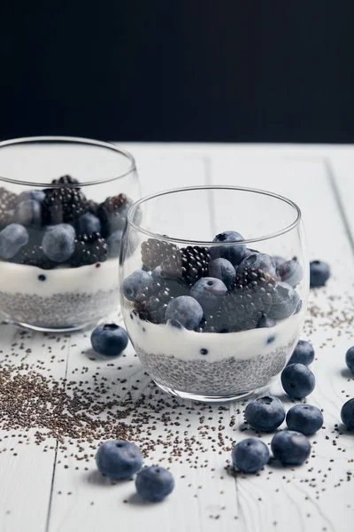 Selective focus of tasty yogurt with chia seeds, blueberries, blackberries near scattered seeds and berries on white wooden isolated on black — Stock Photo