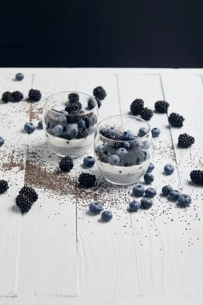 Fuoco selettivo di occhiali con yogurt e bacche vicino a semi di chia sparsi isolati su nero — Stock Photo