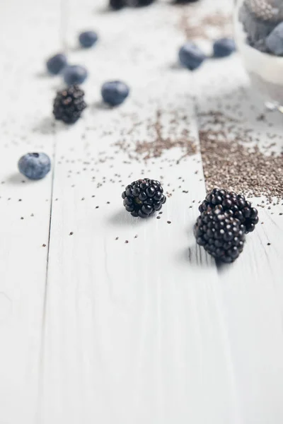 Foco seletivo de amoras silvestres dispersas, mirtilos e sementes de chia na mesa de madeira branca — Fotografia de Stock