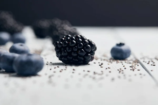 Selective focus of scattered blackberries, blueberries and chia seeds on white wooden table isolated on black — Stock Photo