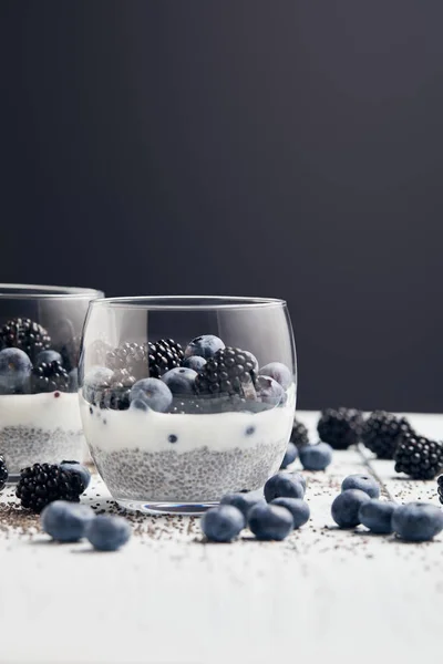 Selektiver Fokus von leckerem Joghurt mit Beeren in der Nähe verstreuter Chiasamen und Beeren auf weißem Holz isoliert auf schwarzem — Stockfoto