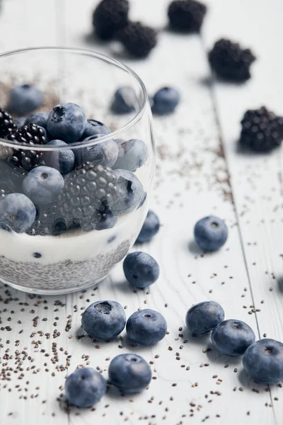 Enfoque selectivo de moras, arándanos y yogur en vidrio cerca de semillas de chía dispersas y bayas en la mesa de madera blanca - foto de stock