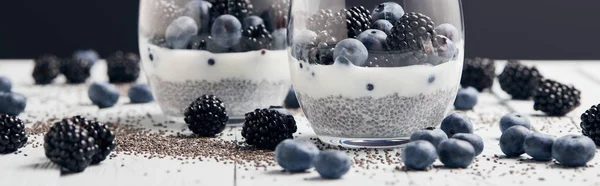 Shot panorámico de yogur con semillas de chía y bayas en vasos cerca de semillas dispersas, arándanos y moras en mesa blanca aislada en negro - foto de stock