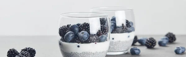 Panoramaaufnahme von leckerem Joghurt mit Chiasamen, Blaubeeren und Brombeeren in Gläsern in der Nähe verstreuter Beeren auf Marmoroberfläche isoliert auf grau — Stockfoto